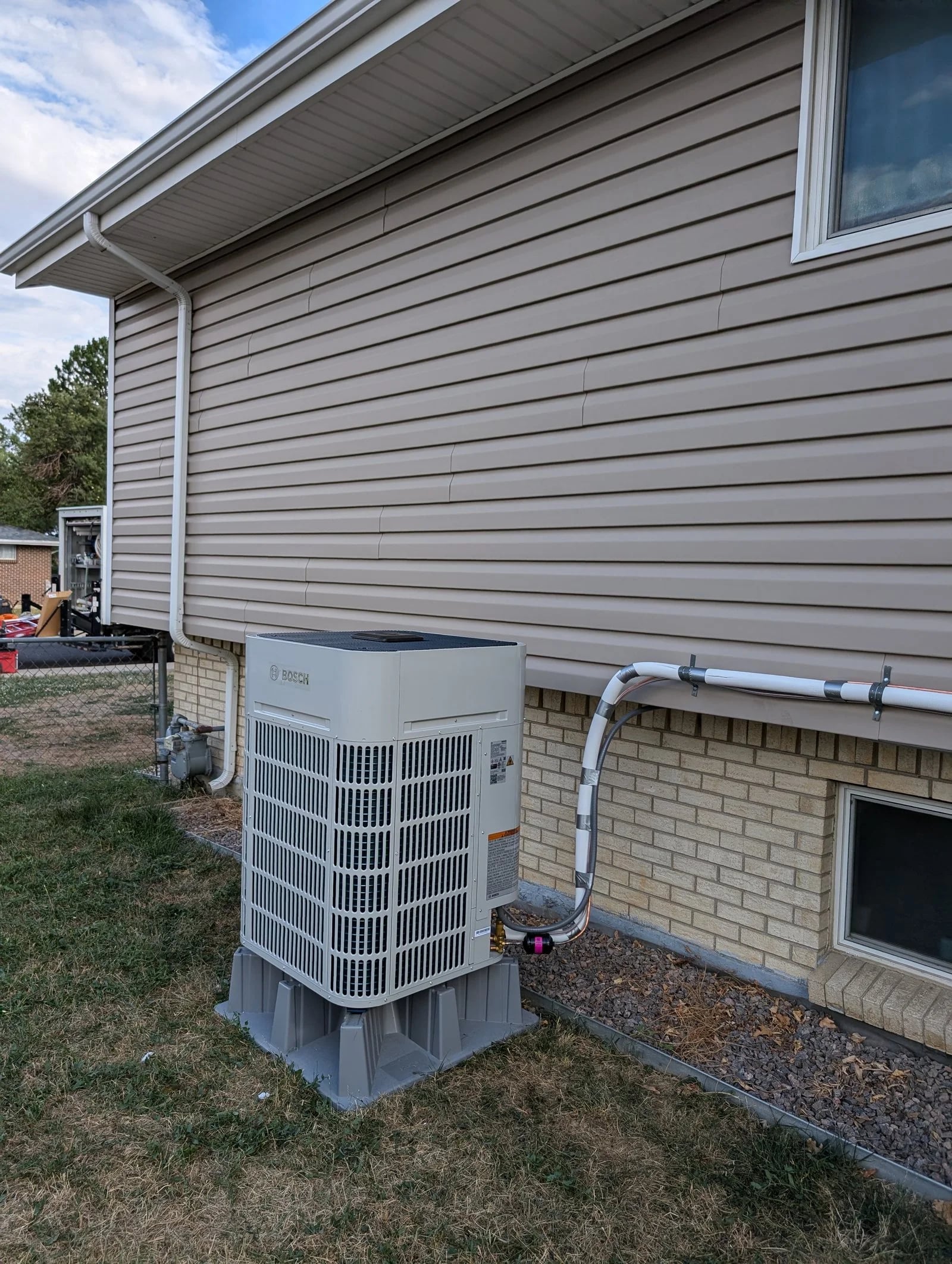 A Bosch outdoor unit on a composite pad on the lawn beside a tan brick and siding house