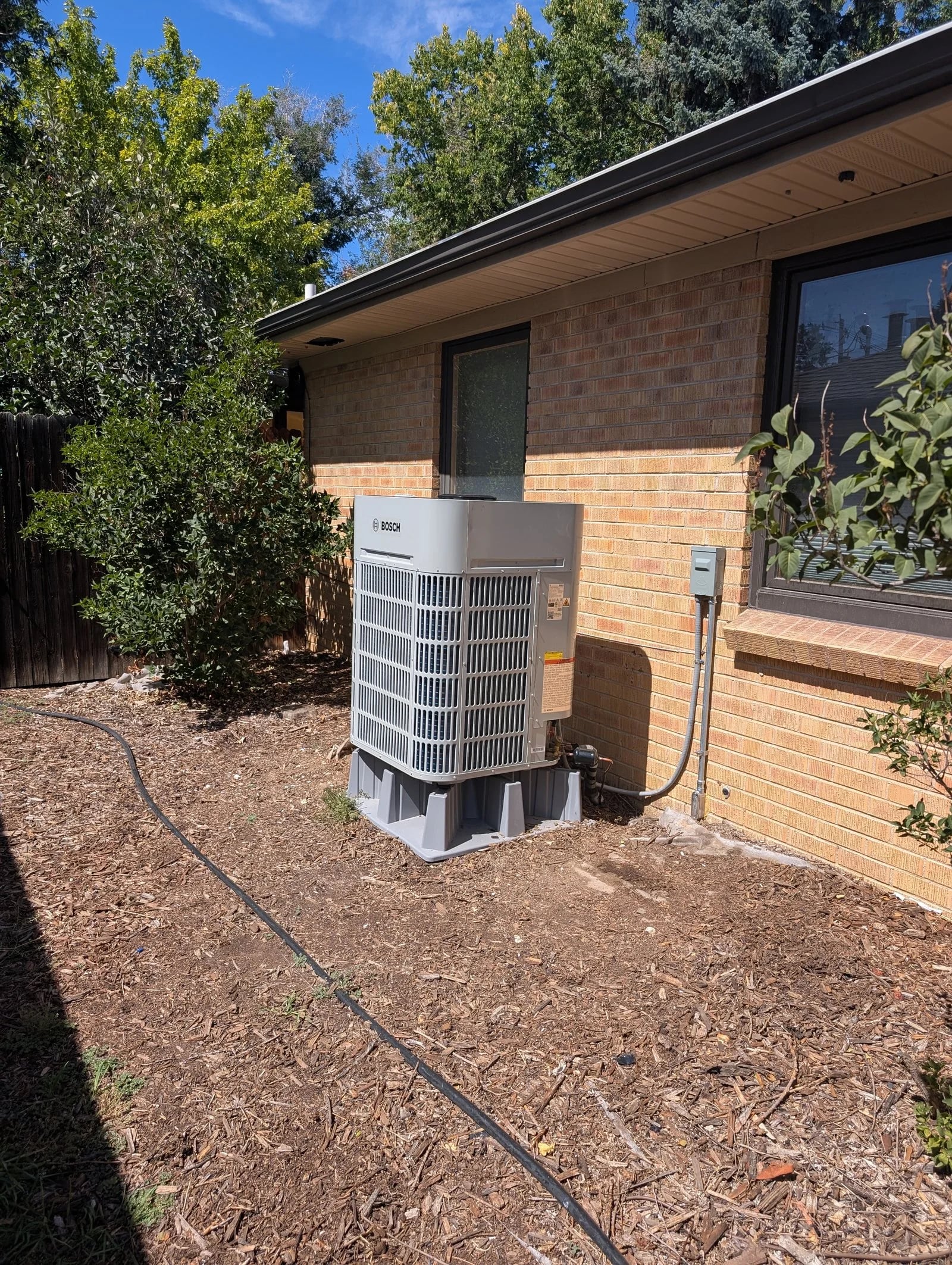 A Bosch outdoor unit on a composite pad beside a brick house with shrubs and a tree
